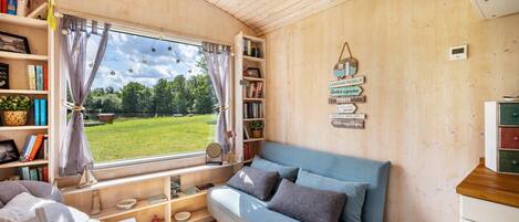 L’intérieur d’un chalet accueillant, avec un canapé bleu, des étagères en bois remplies de livres, une fenêtre avec des rideaux et une vue sur la verdure à l’extérieur.
