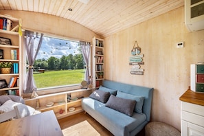 A cozy cabin interior with a blue sofa, wooden shelves with books, a window with curtains, and a view of greenery outside. - Tiny Home 'Mit Blick Auf Den See' with Private Garden, Wi-Fi and Air Conditioning (Fitzen)