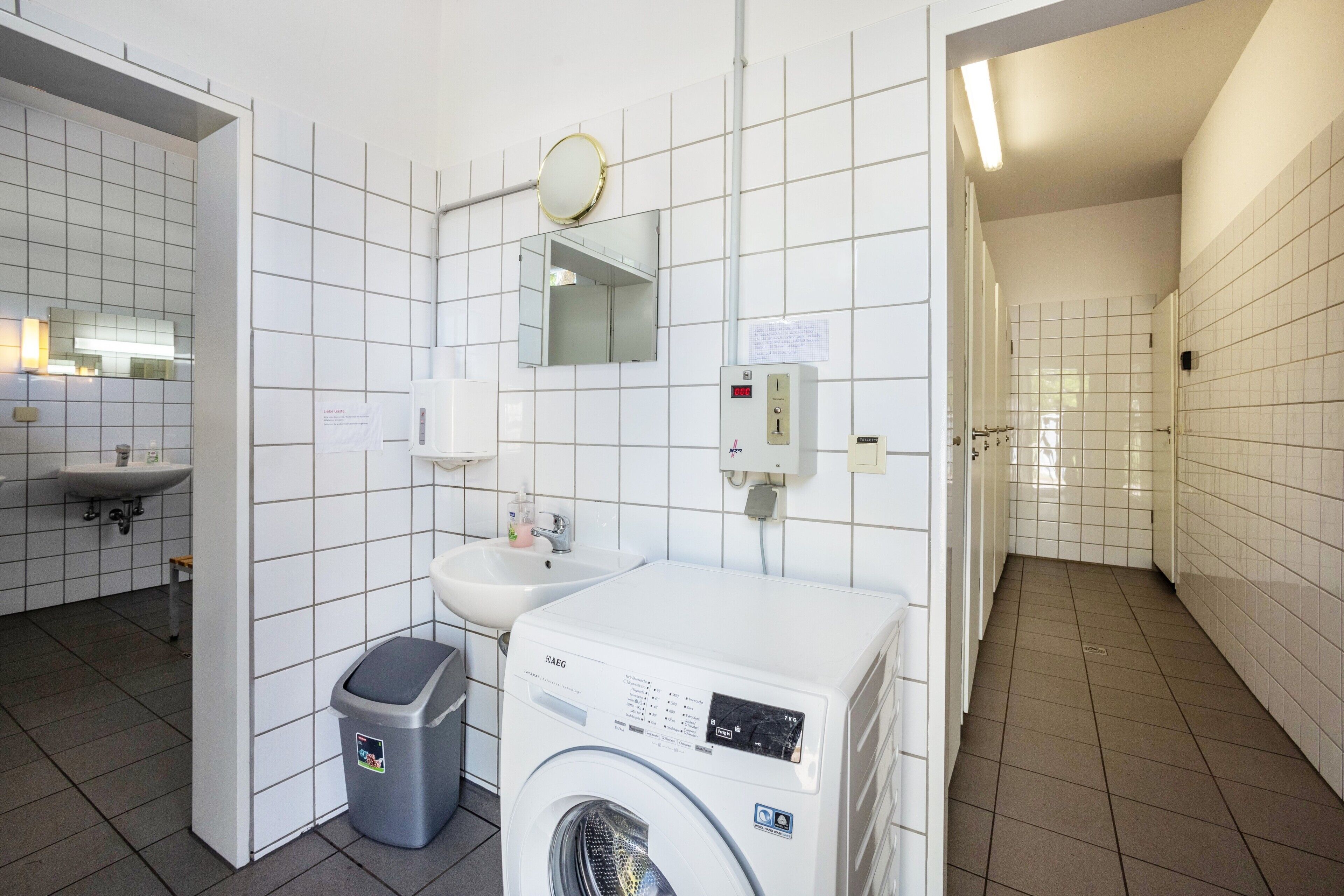 Een openbaar toilet met een wasmachine, een wastafel, een prullenbak en een spiegel.