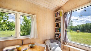 Een knus vertrek met houten inrichting, een eettafel met kommen en lepels, een stoel, een boekenkast, en een groot raam met uitzicht op een gazon.