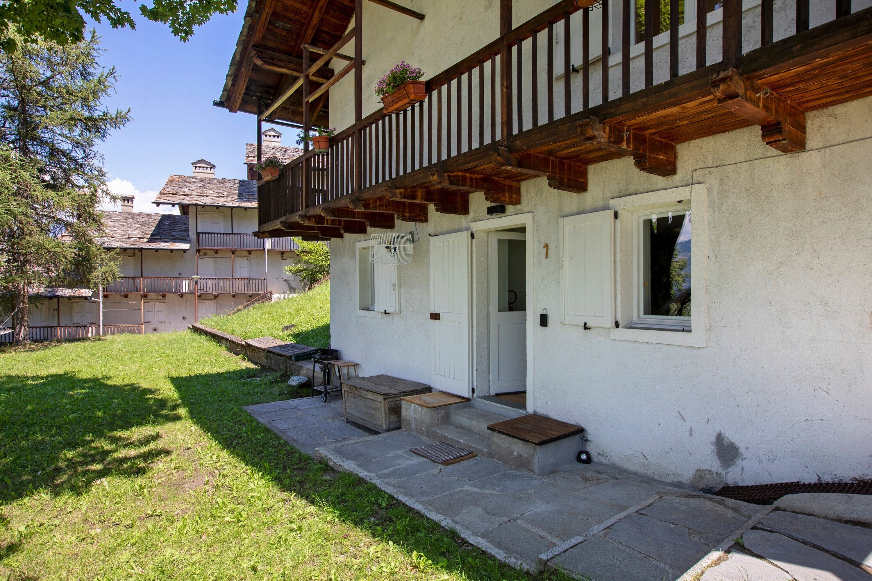 Un chalet traditionnel doté d’un balcon en bois, d’un escalier en pierre et d’un petit espace extérieur aménagé pour s’asseoir.
