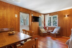 A wooden-paneled room with a dining table, two wicker chairs, a television, and a window with curtains.