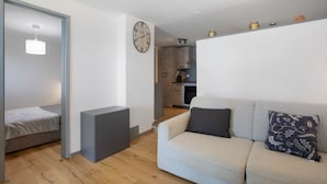 A modern living room with a sofa, a small bed, a wall clock, and a kitchen in the background. - 'Coin Vert' Apartment with Mountain Views, Shared Garden and Wi-Fi (Gressan)