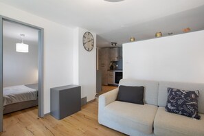 Un salon moderne avec un canapé, un petit lit, une horloge murale et une cuisine en arrière-plan.