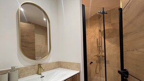 Un baño con lavamanos blanco, grifo dorado y un espejo grande. Cuenta con una ducha con mampara de vidrio y cabezal de ducha dorado.