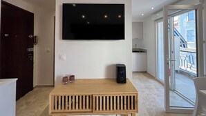 Un salón moderno con un televisor de pantalla plana montado en la pared, una mesa de centro de madera con dos velas, un altavoz negro y una puerta corrediza de vidrio que da a un balcón con vistas a la ciudad.