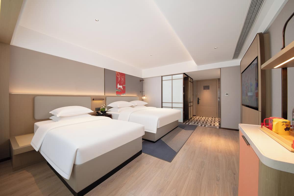 A modern hotel room with two beds, a small seating area, a kitchenette, and a large mirror.