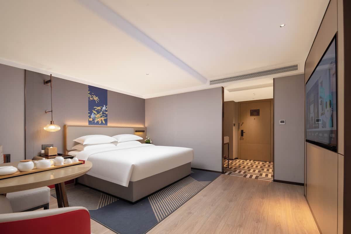A modern hotel room with a large bed, a dining area with a table and chairs, a TV, and a hallway leading to other rooms.