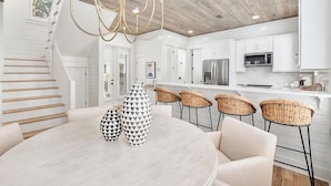 A modern kitchen with white cabinetry, a stainless steel refrigerator, and a microwave. There is a dining area with a round table and four wicker chairs. - Reel McCoy, Seagrove Home with Hot Tub & Fire Pit (Santa Rosa Beach)