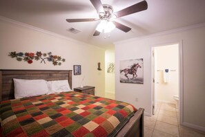 Ein Schlafzimmer mit einem Holzbett, Deckenventilator, einer Quilttdecke, einer an der Wand befestigten Uhr und einer Tür zu einem Badezimmer.