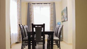 A dining area with a table and chairs, a window with curtains, and a painting on the wall. - Lux Suites Boffar Residence Naivasha (Naivasha)