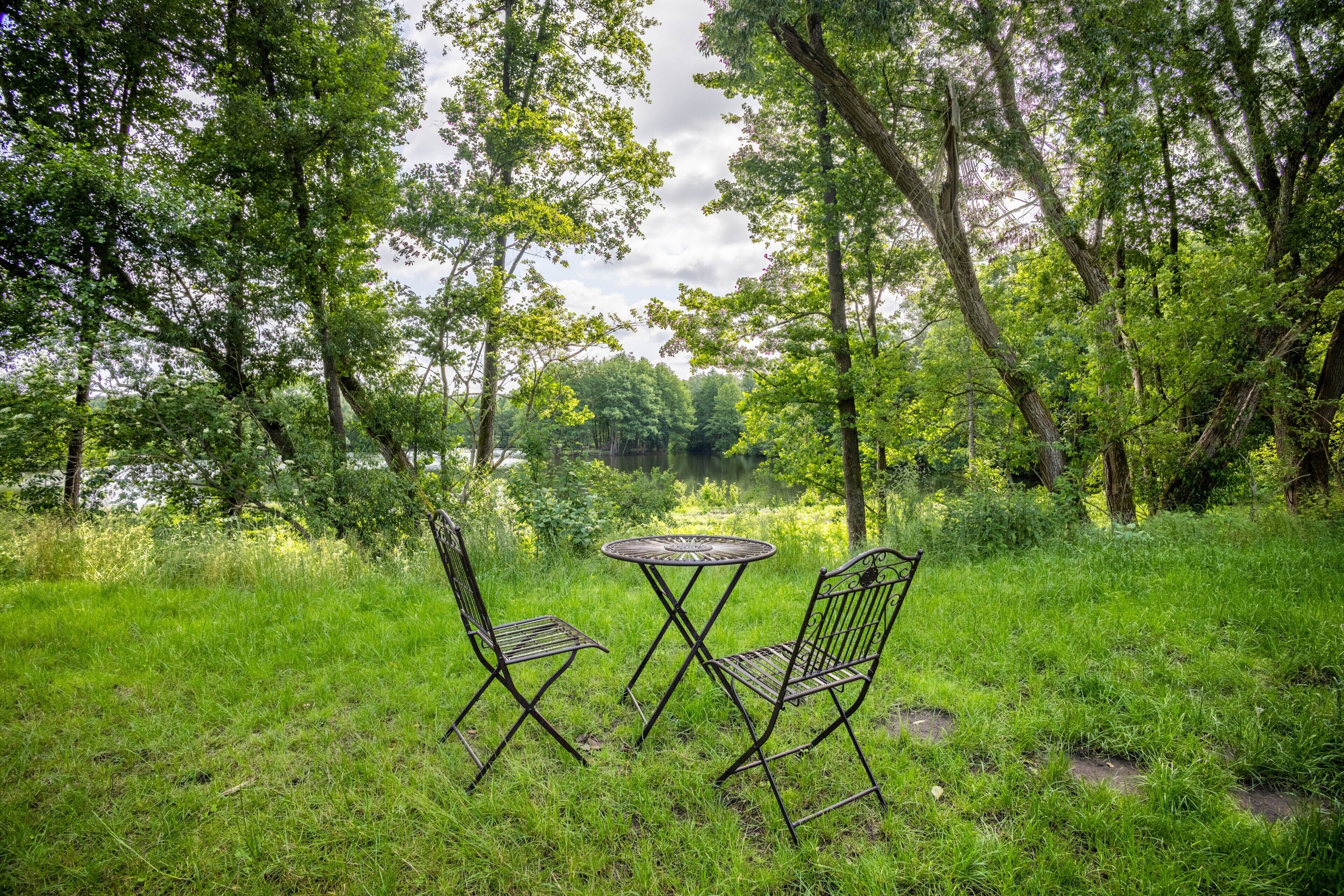 A grassy area with two metal chairs and a round table, surrounded by trees and a body of water.