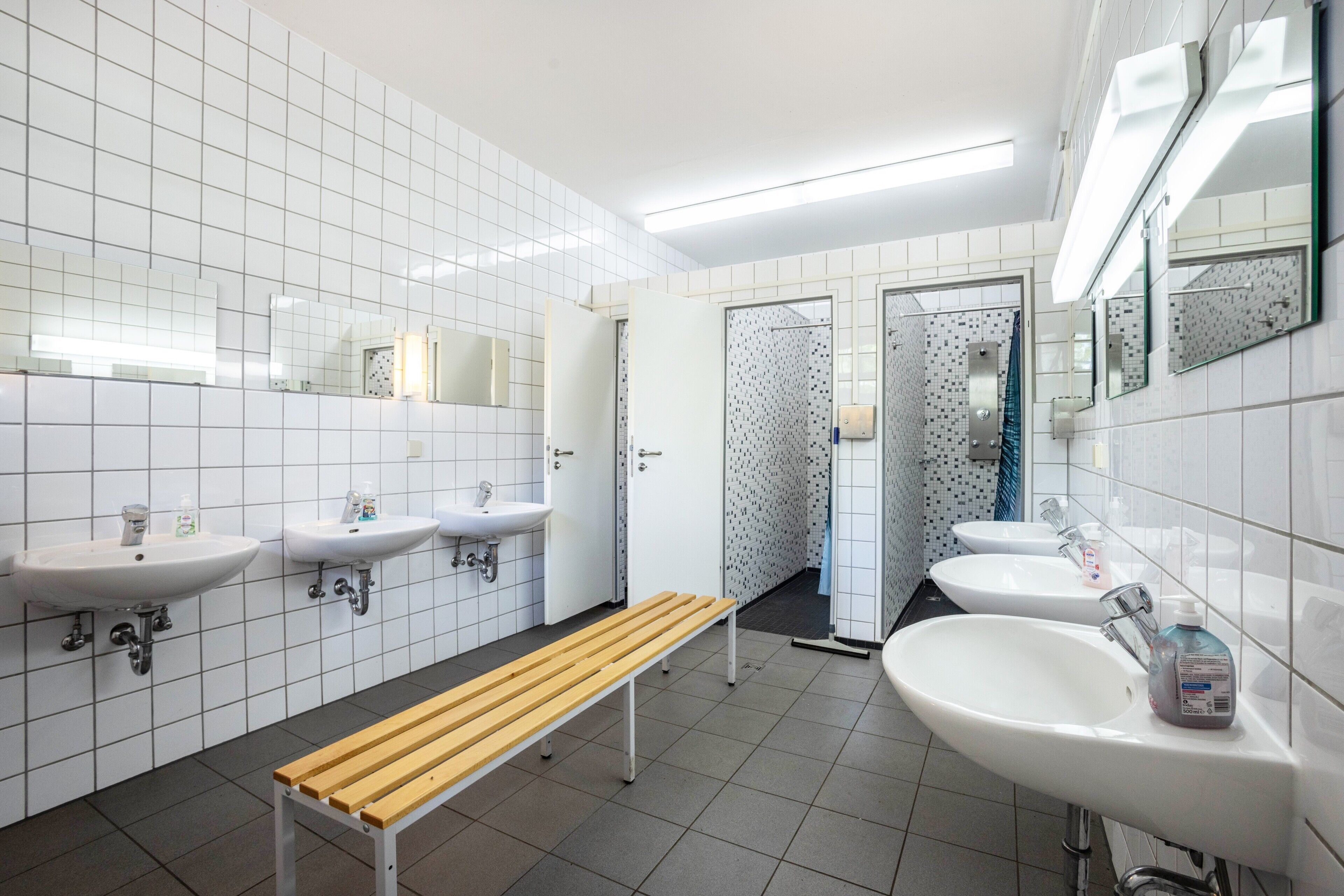 A public restroom with three sinks, a bench, and a door leading to a shower area.