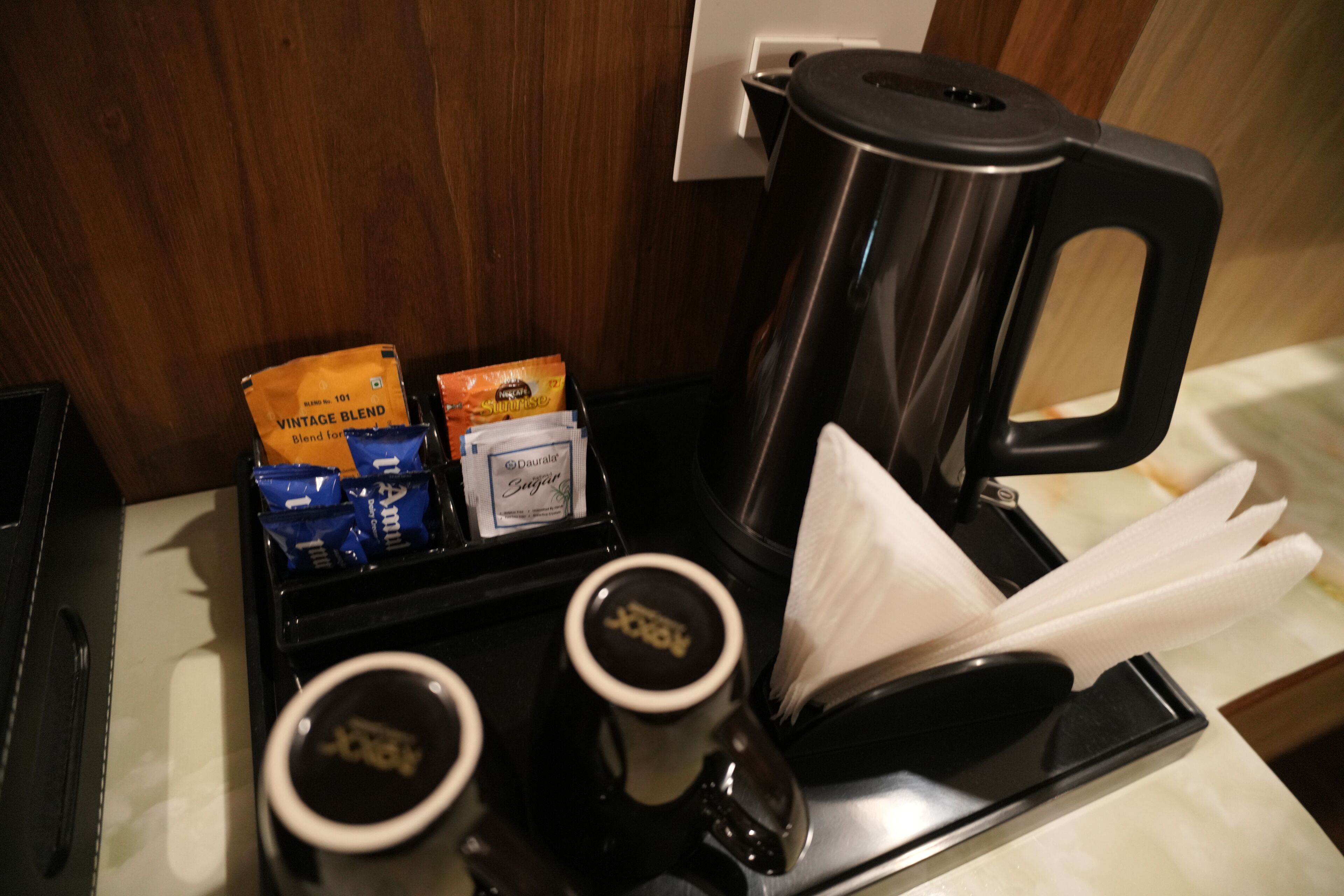 Una estación de café con una cafetera, dos tazas de café, una bandeja con cápsulas de café y un dispensador de servilletas.