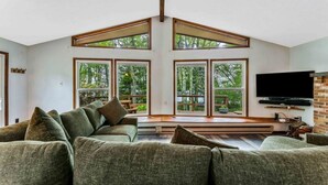 A spacious living room with a large sectional sofa, a flat-screen TV mounted on the wall, and large windows offering a view of the outdoors.