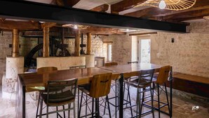 Een rustiek interieur met zichtbare houten balken, een stenen open haard en een lange eettafel met barstoelen.