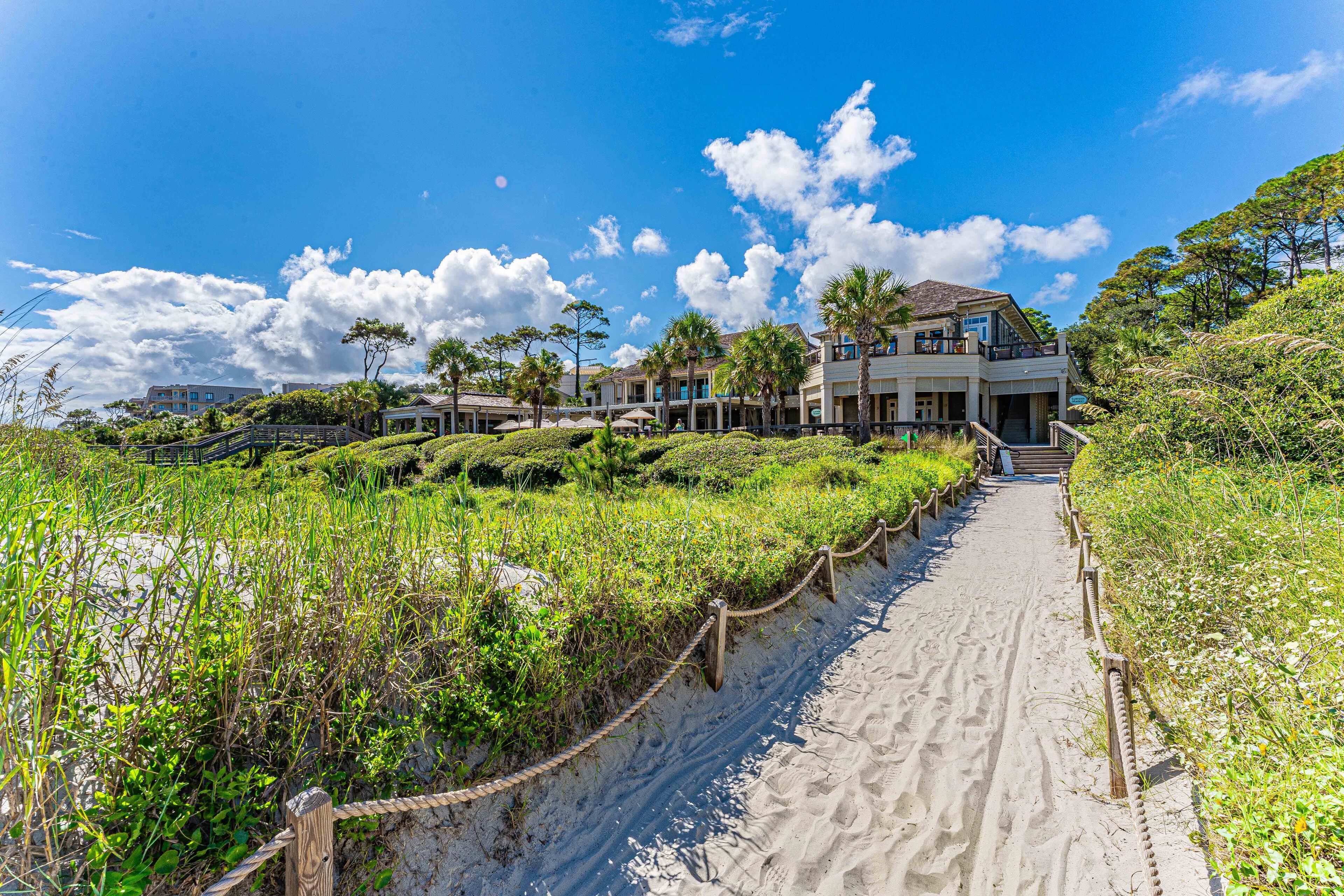 A beachfront property with a sandy pathway leading to a multi-story building, surrounded by lush greenery and palm trees.
