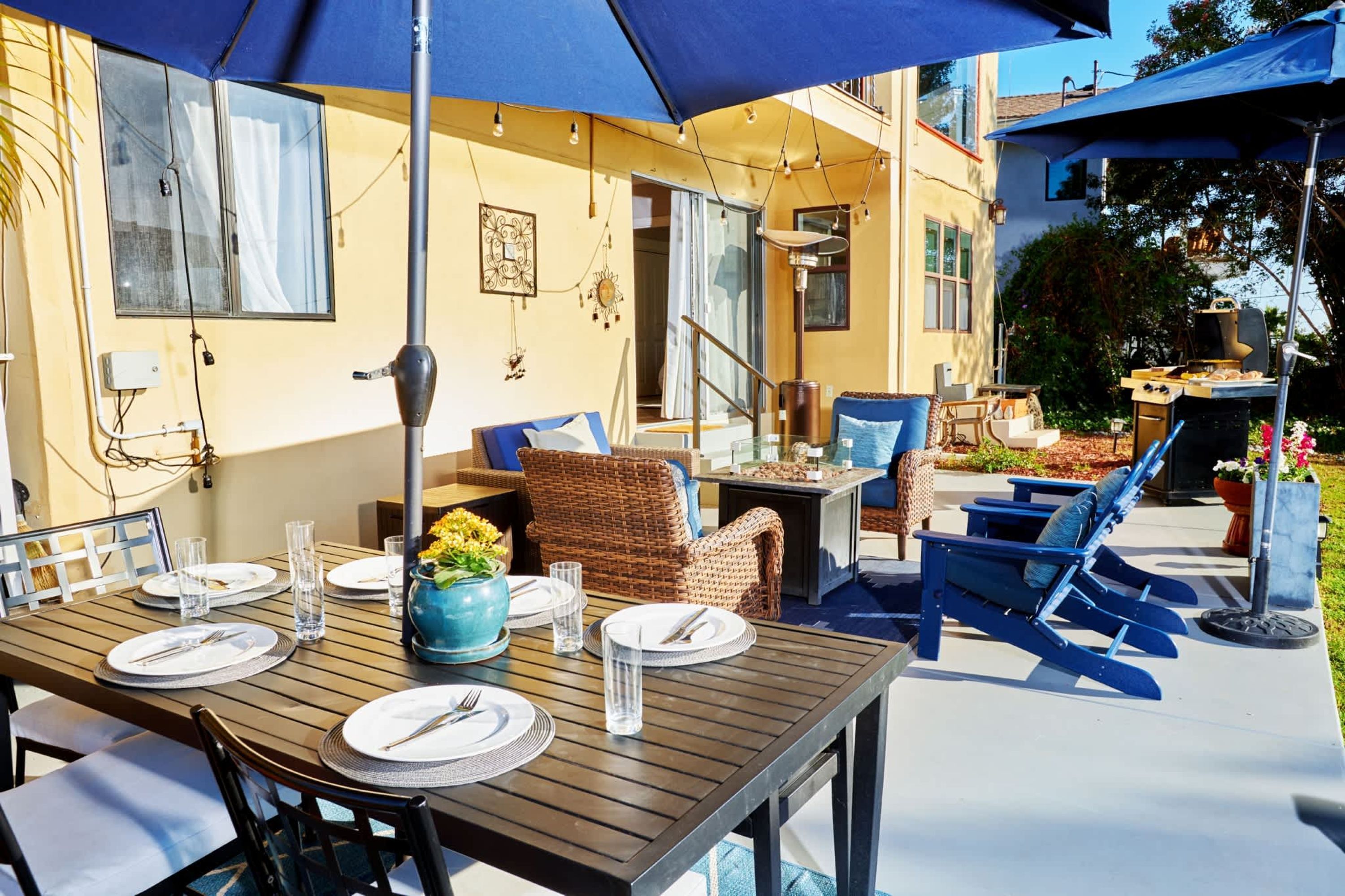 Un espace de restauration en plein air avec une table en bois disposée pour quatre personnes, des chaises bleues et un parasol bleu offrant de l’ombre.