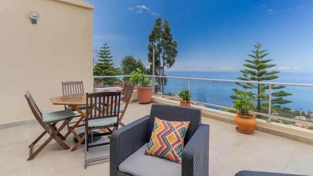 Ein Balkon mit Tisch und Stühlen, Blumentöpfen und Blick auf das Meer.