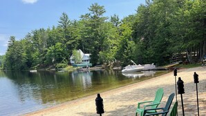 A lakeside setting with two Adirondack chairs, a fire pit, and a boat docked near a house.