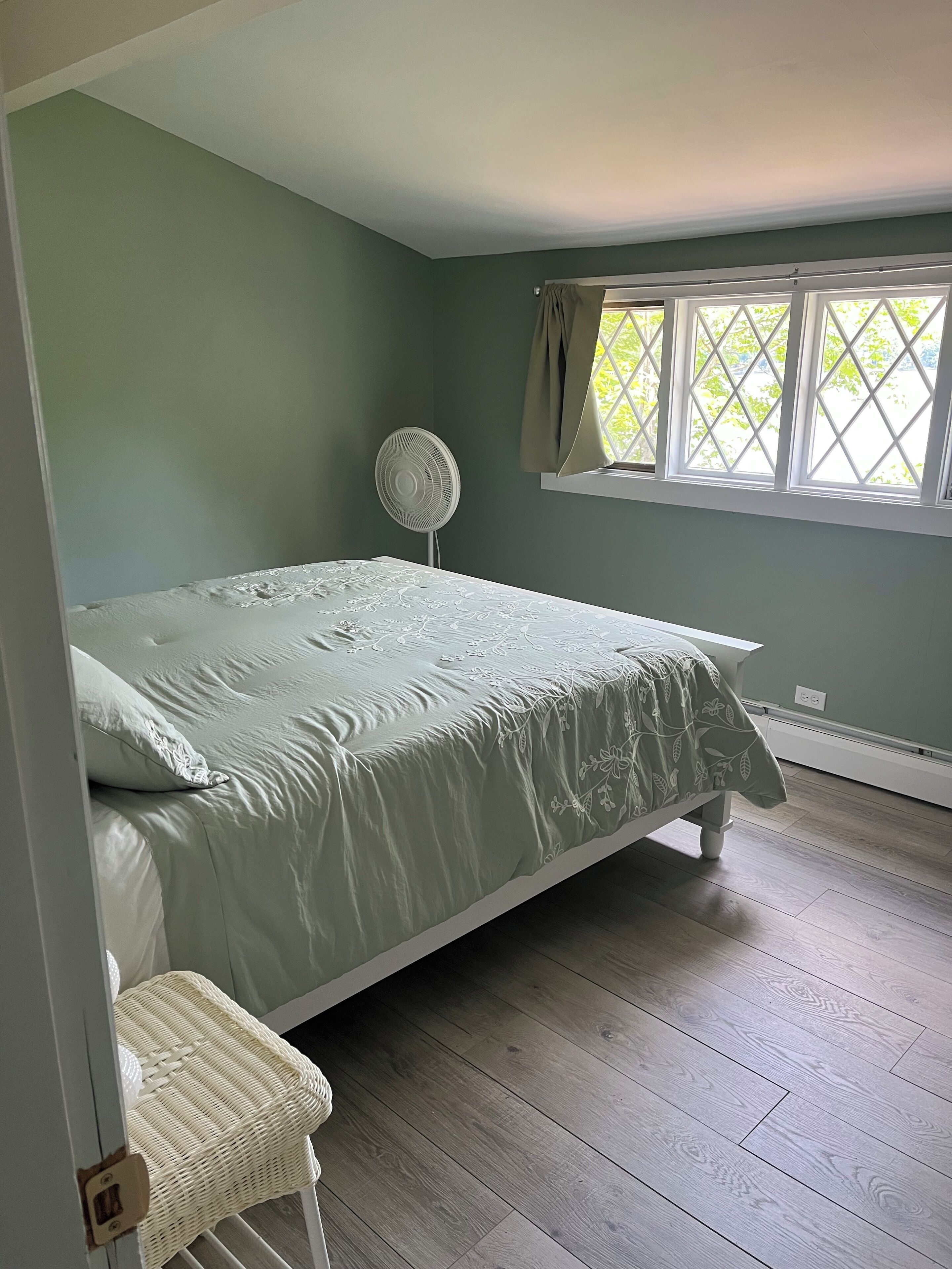 Een slaapkamer met een bed, een ventilator, een raam met gordijnen en een rieten stoel.