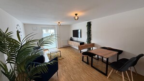 Ett modernt vardagsrum med trägolv, en blå soffa, ett trämatbord med stolar, en tv som är monterad på väggen och en stor växt i hörnet.