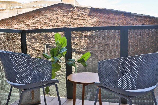 Un balcón con dos sillas de metal y una mesita pequeña, una planta en maceta y vista a un muro de ladrillo.