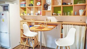 Une cuisine de taille réduite, avec des étagères en bois, un réfrigérateur et deux chaises blanches.