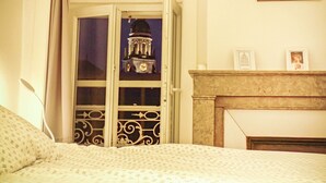 Une pièce avec un lit, une cheminée, un clocher visible par la fenêtre et un tableau encadré au mur.