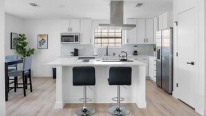 A modern kitchen with white cabinets, a central island with bar stools, stainless steel appliances, and a window with blinds. - The Belvedere Pet Friendly Spacious Yard (Plano)