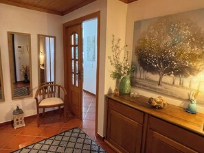 Un pasillo con una silla de madera, piso de baldosas, una puerta de madera y un cuadro de un árbol.