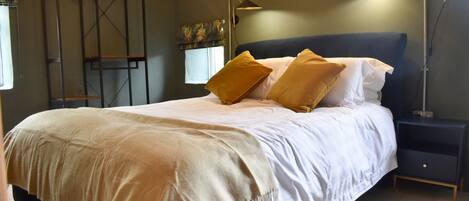 A neatly made bed with a white and beige duvet, two mustard-colored pillows, and a dark headboard. There is a bedside table with a lamp and a small shelf. A metal shelving unit with clothes hangs on the wall to the left.