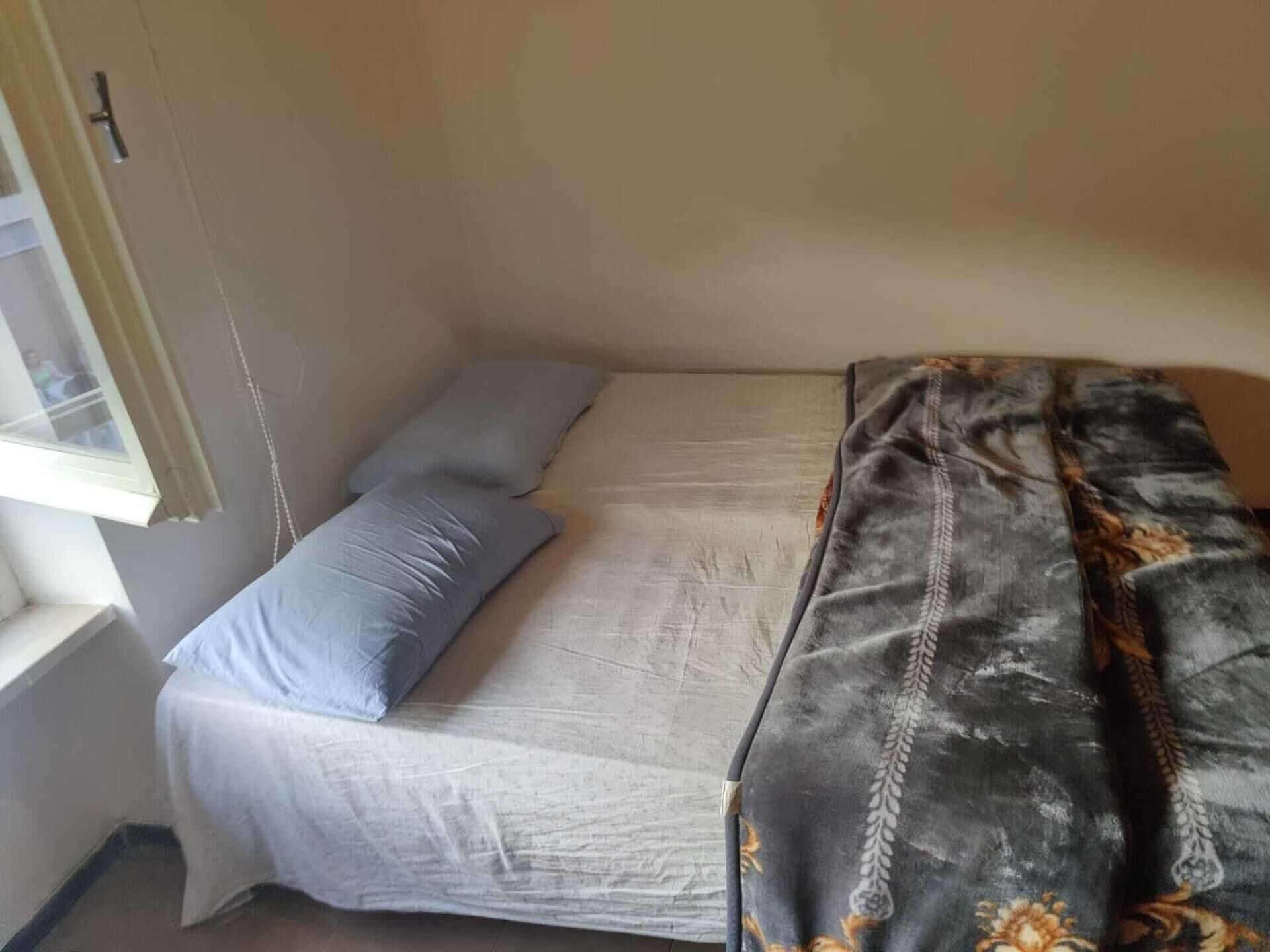 A small, single-bed room with a window, a bedside table, and a patterned blanket.