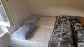 A small, single-bed room with a window, a bedside table, and a patterned blanket.