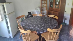 Un tavolo da pranzo rotondo con tovaglia in marmo bianco e nero, circondato da quattro sedie in legno. A sinistra si trova un frigorifero bianco, e a destra un mobiletto in legno con ante in vetro. Un ventilatore è posizionato a terra vicino al tavolo.