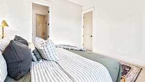 A bedroom with two beds, a bedside table, a lamp, and a door leading to another room. - Dark & Sleek 3BR Escape by the Golf Course (Boise)