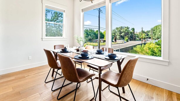 A dining area with a table set for four, large windows, and a view of greenery and a road. - Dark & Sleek 3BR Escape by the Golf Course (Boise)