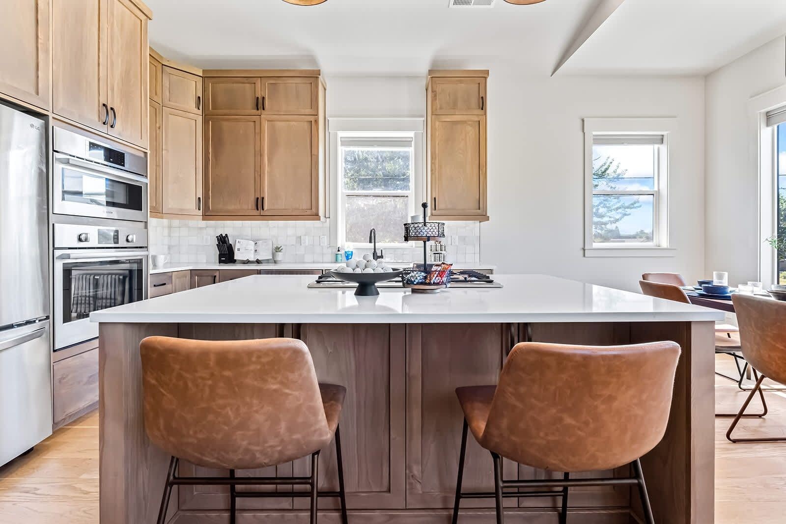A modern kitchen with wooden cabinets, a large island with bar stools, stainless steel appliances, and a view through the window.