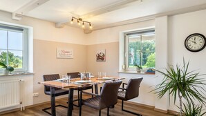 A dining room with a wooden table, four chairs, a clock on the wall, and a window with a view of greenery. - Apartment 'Mayerhofer 1' with Private Terrace, Shared Garden and Wi-Fi (Arnstorf)
