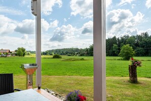 A covered outdoor area with a grill, a table, and a view of a green field and trees. - Apartment 'Mayerhofer 1' with Private Terrace, Shared Garden and Wi-Fi (Arnstorf)