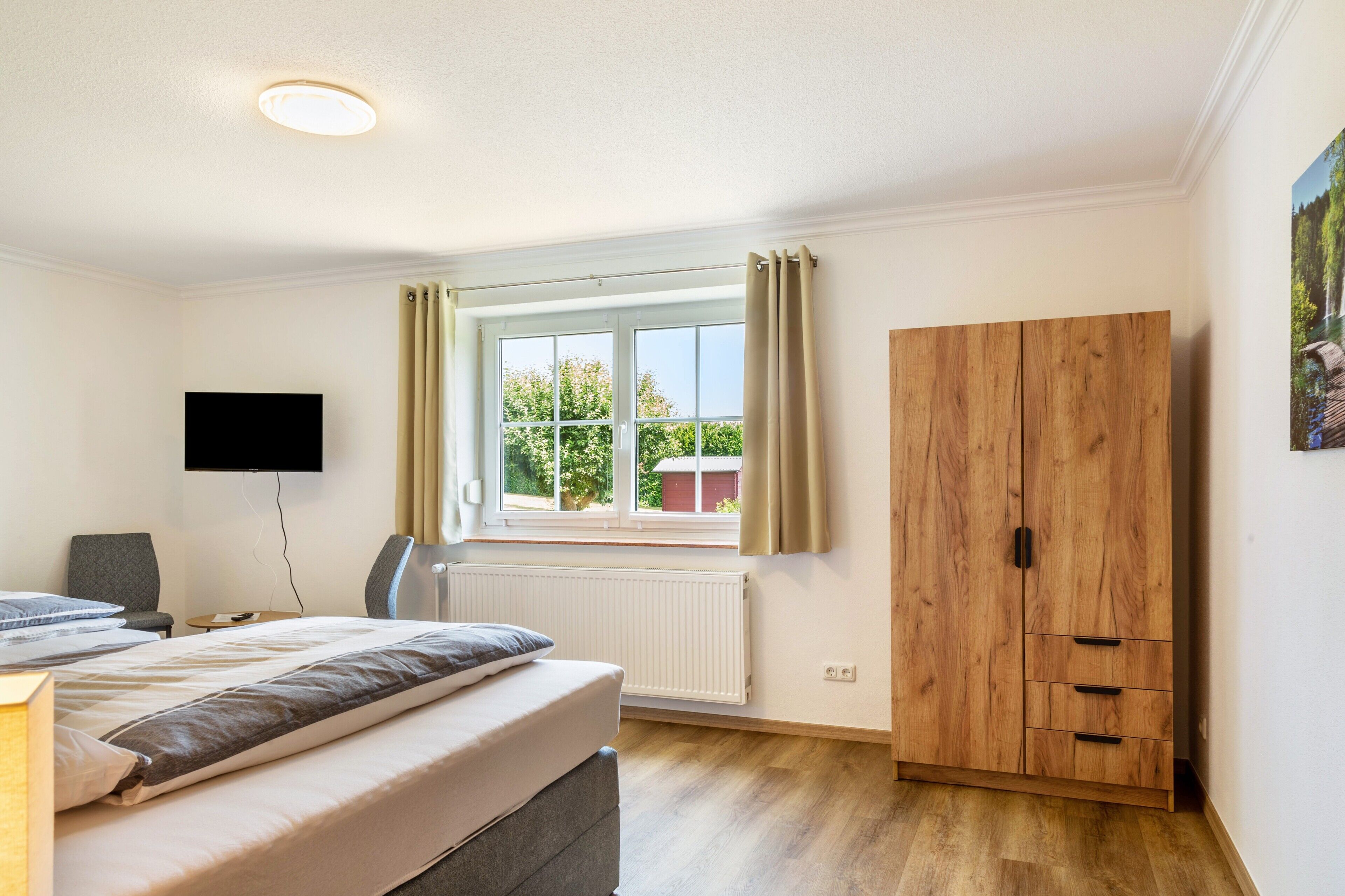 Ein Schlafzimmer mit einem Bett, einem Fernseher, einem Fenster mit Vorhängen, einem Holzschrank und einer Heizung.