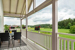 Een overdekt buitengebied met een tafel en stoelen, een witte balustrade en uitzicht op een groen landschap.