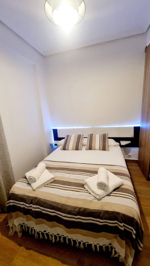 Een netjes opgemaakt bed met gestreept beddengoed en opgerolde handdoeken, een nachtkastje met een lamp, en een moderne plafondlamp.