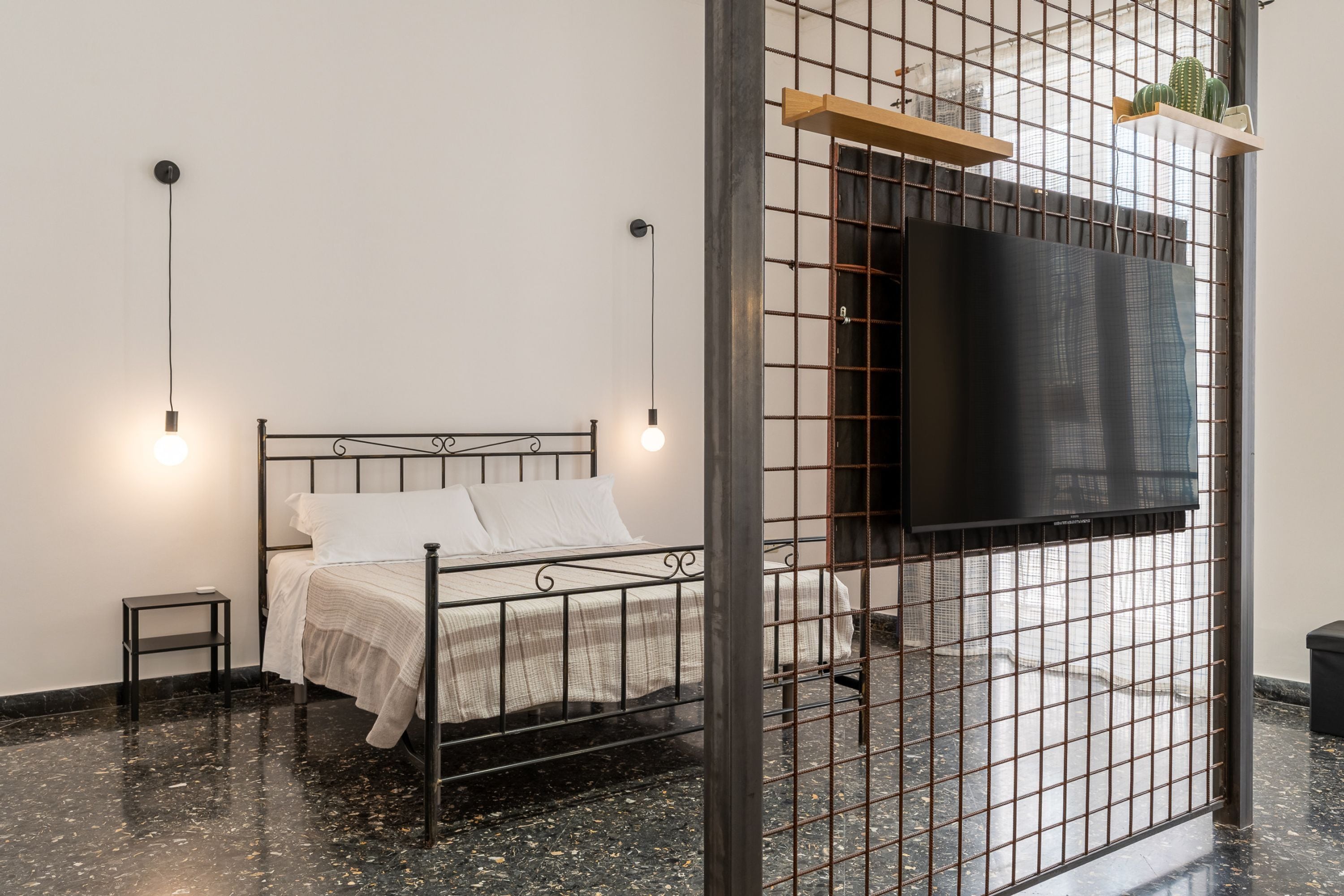 Une chambre moderne avec un lit en métal, une petite table de chevet, un téléviseur fixé sur un mur en treillis et une fenêtre donnant sur la ville.