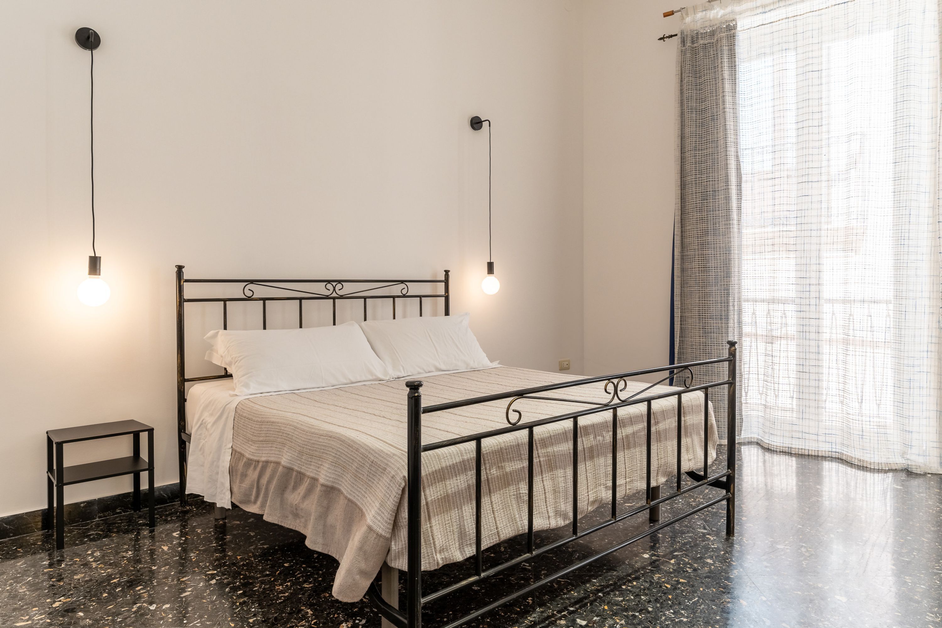 Quarto com estrutura de cama de metal, roupa de cama branca, uma pequena mesa de cabeceira e duas luminárias fixadas na parede.
