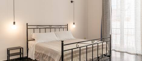 Quarto com estrutura de cama de metal, roupa de cama branca, uma pequena mesa de cabeceira e duas luminárias fixadas na parede.