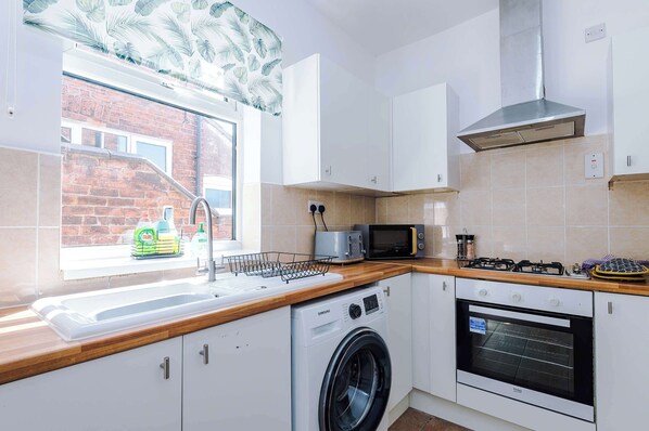 A compact kitchen with a washing machine, microwave, and oven, a window with a view of a brick building, and a tiled backsplash.