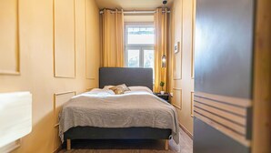 Ein schmales Hotelzimmer mit einem Bett, einem Nachttisch, einem Fenster mit Vorhängen und einem Holzboden.