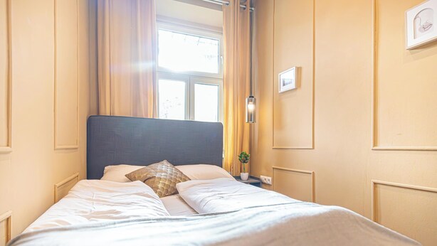 Ein ordentlich bezogenes Bett mit Kopfteil, ein Nachttisch mit Lampe und ein Fenster mit Vorhängen.