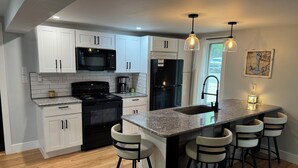 A modern kitchen with white cabinets, a black stove, a black refrigerator, and a granite countertop. There are four bar stools and pendant lights above the island.